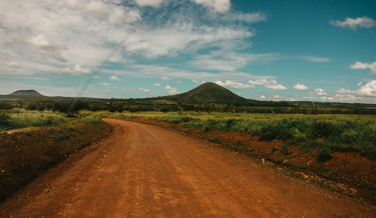 hero-img-02 Explore the serene beauty of a rural dirt road winding through the lush Tanzanian countryside under a bright sky.
